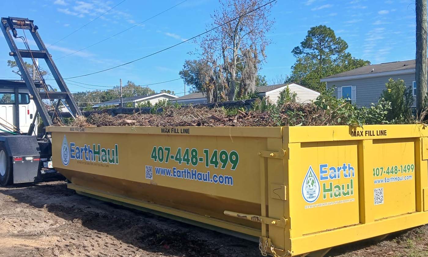 Branches, brush, and a tree trunk on the ground near a roll-off dumpster in Melbourne Beach, FL.